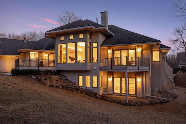 back of property at dusk featuring roof with shingles, a chimney, and stucco siding