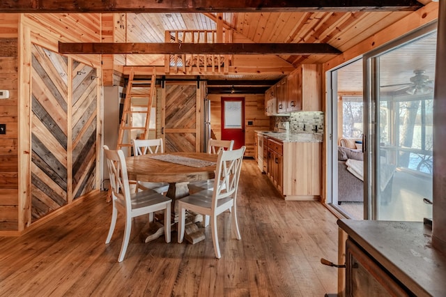 dining room featuring ceiling fan, beam ceiling, wooden walls, light hardwood / wood-style floors, and a barn door