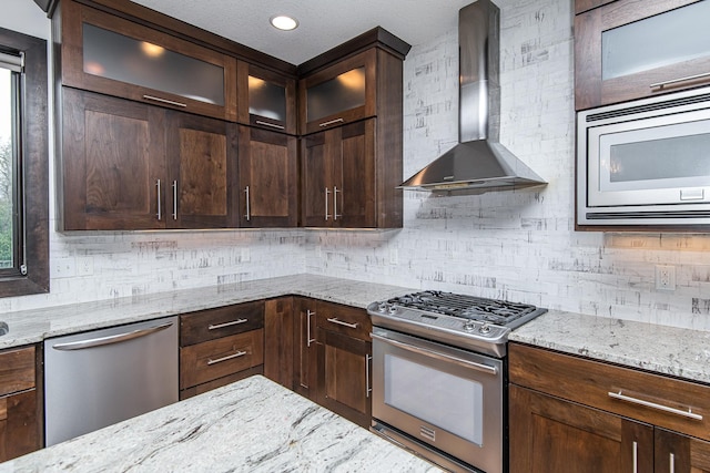 kitchen with light stone counters, wall chimney range hood, decorative backsplash, and appliances with stainless steel finishes