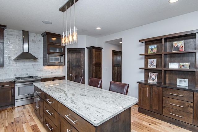 kitchen featuring wall chimney range hood, hanging light fixtures, stainless steel appliances, a center island, and light stone counters