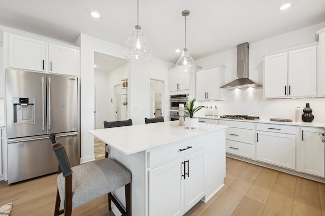 kitchen featuring wall chimney exhaust hood, a breakfast bar, appliances with stainless steel finishes, pendant lighting, and white cabinets