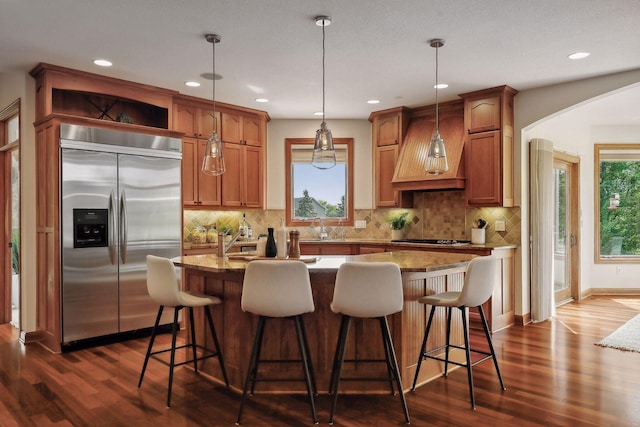 kitchen with custom exhaust hood, built in refrigerator, decorative light fixtures, light stone countertops, and a kitchen island with sink