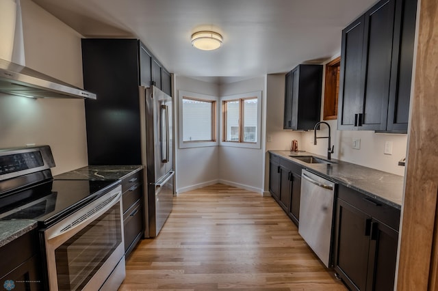 kitchen with sink, wall chimney range hood, appliances with stainless steel finishes, dark stone countertops, and light hardwood / wood-style floors