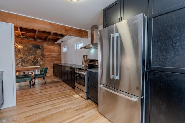 kitchen with wooden walls, beamed ceiling, stainless steel appliances, light hardwood / wood-style floors, and wall chimney range hood