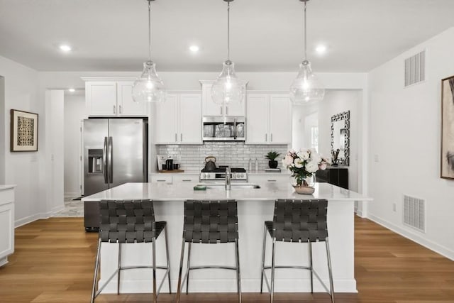 kitchen with hanging light fixtures, a center island with sink, stainless steel appliances, decorative backsplash, and white cabinets