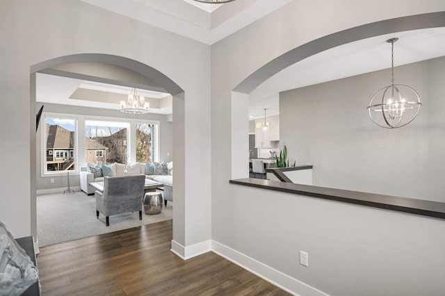 hallway with a chandelier, arched walkways, baseboards, dark wood-style floors, and a raised ceiling