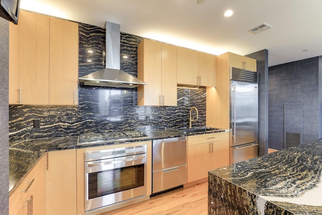 kitchen featuring light brown cabinetry, sink, dark stone countertops, stainless steel appliances, and wall chimney exhaust hood