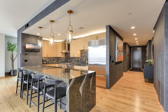 kitchen featuring wall chimney exhaust hood, light brown cabinetry, built in fridge, pendant lighting, and dark stone counters