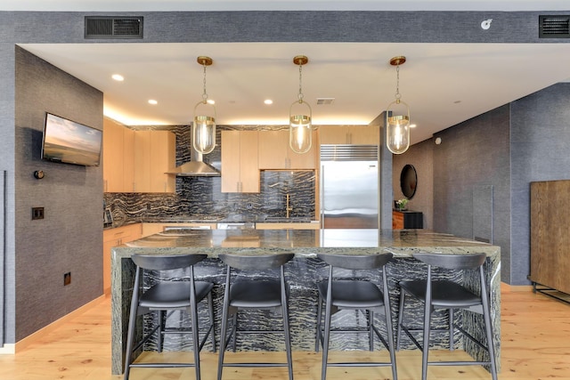 kitchen with wall chimney exhaust hood, light brown cabinetry, built in refrigerator, hanging light fixtures, and light wood-type flooring