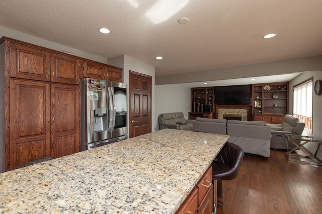 kitchen featuring dark hardwood / wood-style flooring, a kitchen bar, light stone counters, stainless steel fridge with ice dispenser, and a brick fireplace