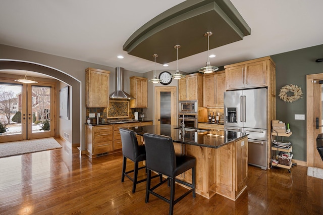 kitchen with wall chimney range hood, hanging light fixtures, stainless steel appliances, a center island with sink, and decorative backsplash