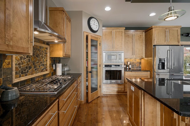 kitchen featuring wall chimney exhaust hood, wood-type flooring, dark stone counters, pendant lighting, and stainless steel appliances