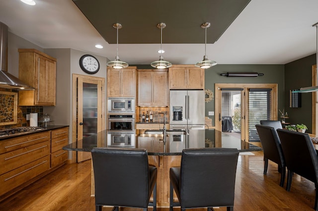 kitchen with wall chimney exhaust hood, sink, hanging light fixtures, a center island with sink, and stainless steel appliances