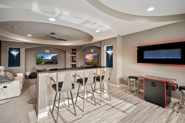 cinema room with ceiling fan, bar area, a tray ceiling, and light wood-type flooring