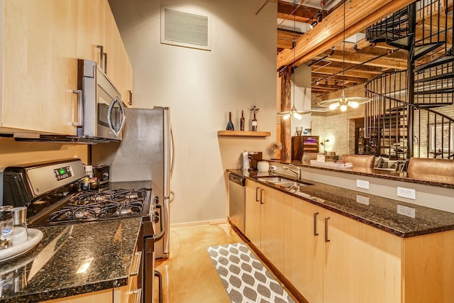 kitchen featuring sink, appliances with stainless steel finishes, dark stone countertops, light brown cabinetry, and decorative light fixtures