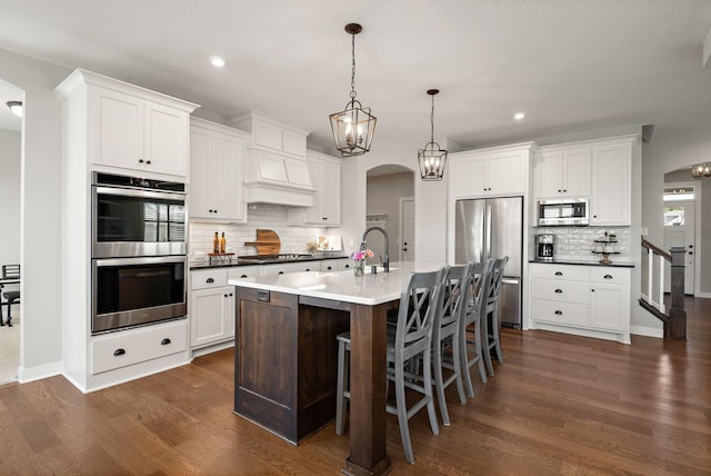 kitchen with appliances with stainless steel finishes, a kitchen island with sink, white cabinets, and decorative light fixtures