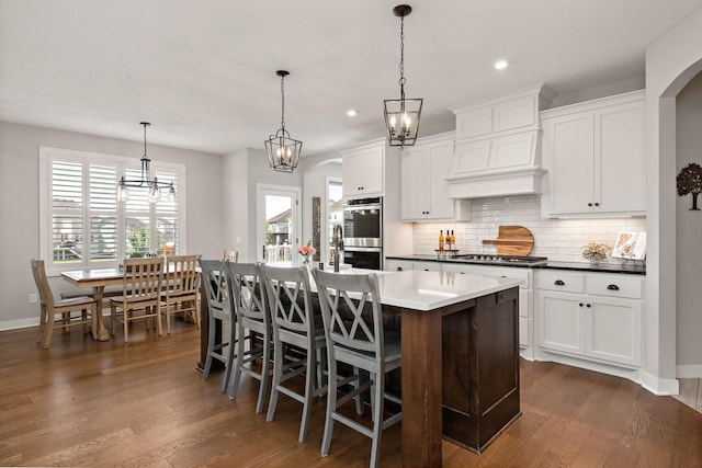 kitchen featuring hanging light fixtures, backsplash, stainless steel appliances, white cabinets, and a center island with sink