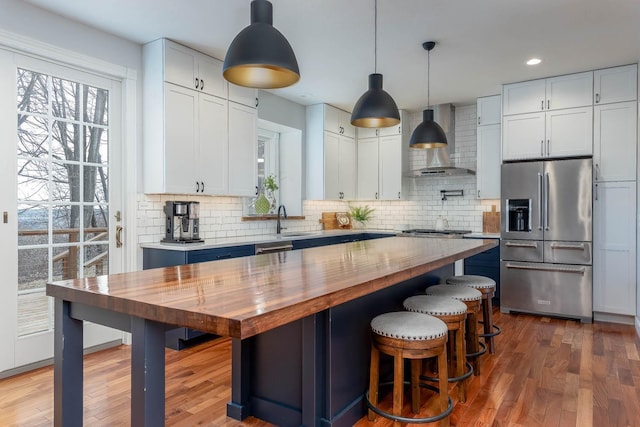 kitchen with butcher block countertops, wall chimney range hood, appliances with stainless steel finishes, white cabinetry, and a kitchen island