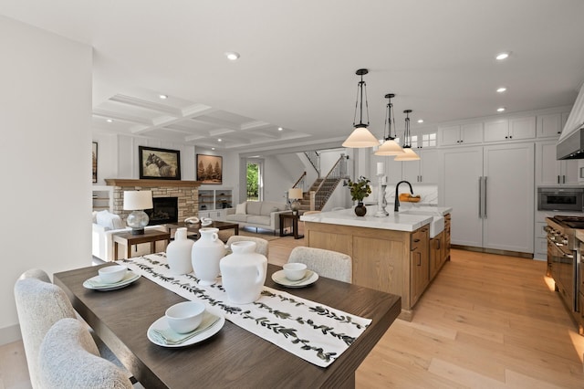 dining area featuring a stone fireplace, beamed ceiling, sink, coffered ceiling, and light hardwood / wood-style floors