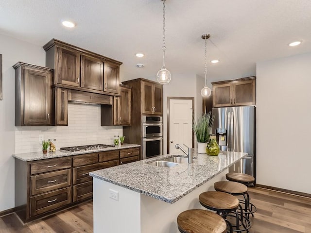 kitchen with sink, light stone counters, an island with sink, pendant lighting, and stainless steel appliances