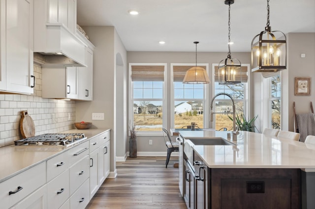 kitchen with white cabinetry, hanging light fixtures, stainless steel gas stovetop, a kitchen island with sink, and backsplash