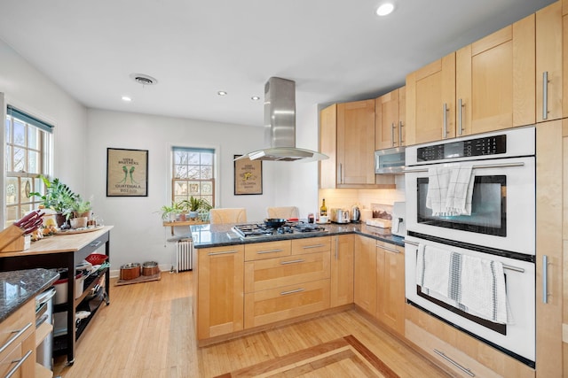 kitchen with radiator heating unit, double oven, stainless steel gas stovetop, island exhaust hood, and kitchen peninsula