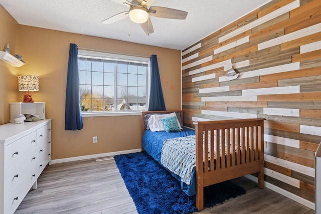 bedroom with light wood finished floors, wooden walls, an accent wall, baseboards, and a textured ceiling