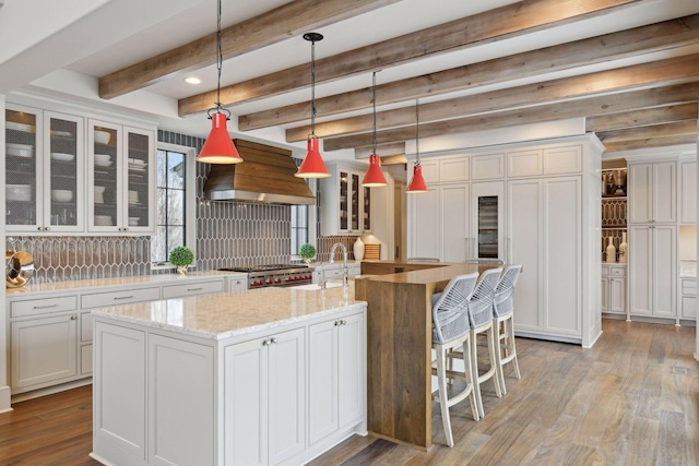 kitchen with custom exhaust hood, white cabinetry, hanging light fixtures, and a kitchen island with sink