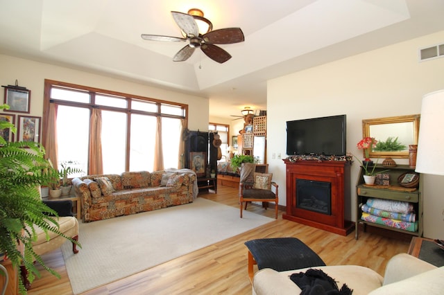 living area featuring wood finished floors, visible vents, a ceiling fan, a tray ceiling, and a glass covered fireplace