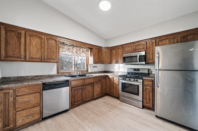 kitchen with light wood-style flooring, a sink, vaulted ceiling, appliances with stainless steel finishes, and dark countertops