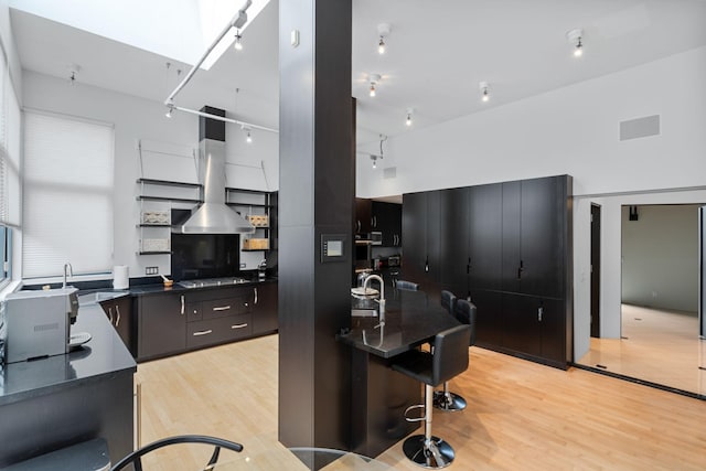 kitchen with a sink, wall chimney range hood, light wood-type flooring, dark countertops, and cooktop