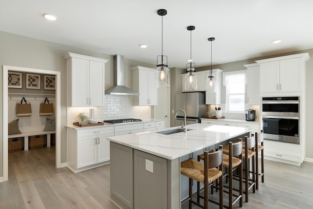 kitchen featuring wall chimney exhaust hood, sink, appliances with stainless steel finishes, an island with sink, and white cabinets