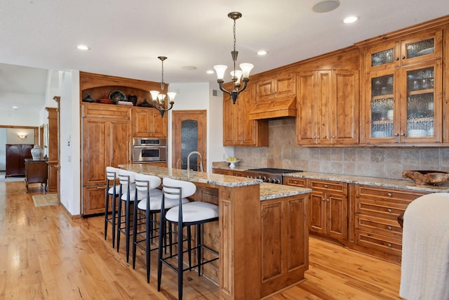 kitchen with custom exhaust hood, light stone counters, a center island with sink, pendant lighting, and stainless steel appliances