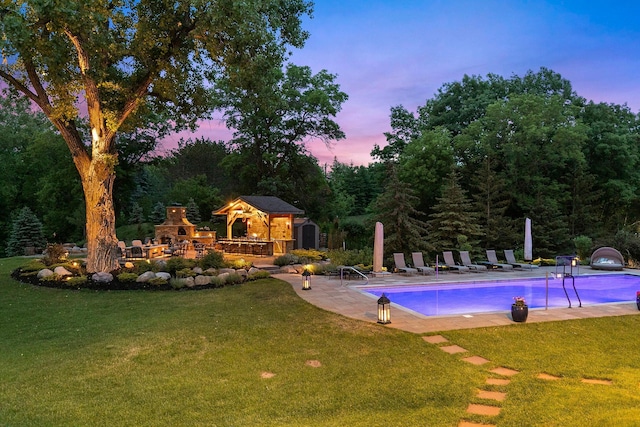 pool at dusk with a lawn, an outdoor structure, a patio, and an outdoor fireplace