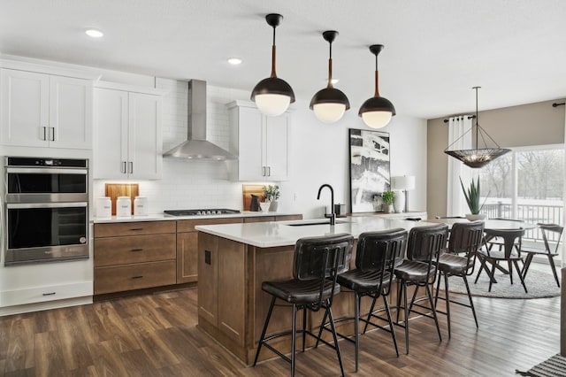 kitchen featuring sink, wall chimney range hood, an island with sink, stainless steel appliances, and white cabinets