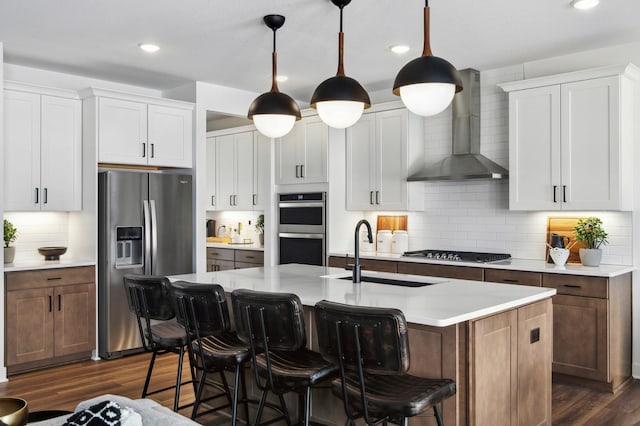 kitchen featuring white cabinetry, decorative light fixtures, stainless steel appliances, a kitchen island with sink, and wall chimney range hood