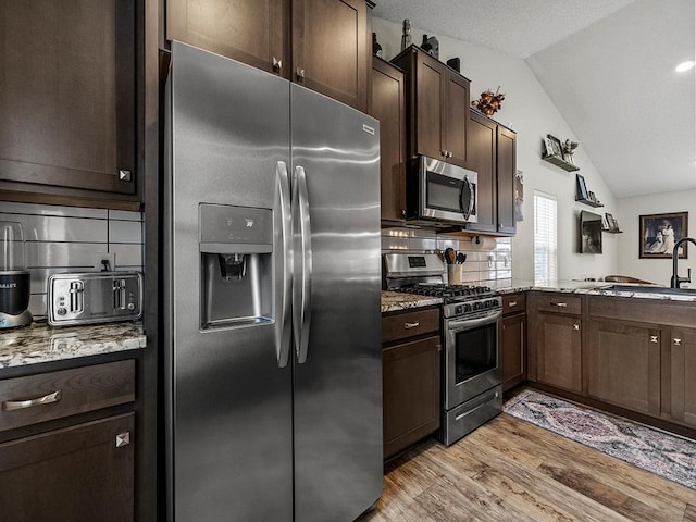 kitchen featuring appliances with stainless steel finishes, light stone counters, dark brown cabinetry, light hardwood / wood-style floors, and vaulted ceiling