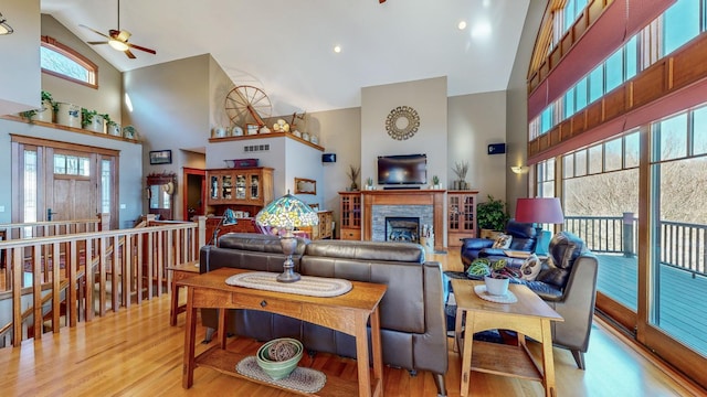 living room with ceiling fan, a stone fireplace, high vaulted ceiling, and light hardwood / wood-style flooring