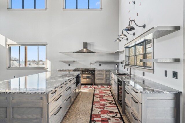 kitchen with a sink, open shelves, a high ceiling, high end stove, and wall chimney exhaust hood