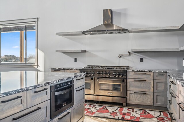 kitchen featuring double oven range, light stone counters, open shelves, and wall chimney exhaust hood