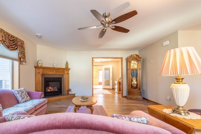 living area featuring wood finished floors, a glass covered fireplace, ceiling fan, and baseboards
