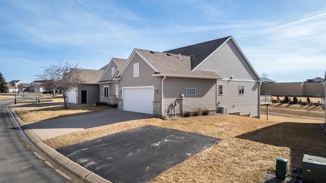 view of side of home featuring a shingled roof, an attached garage, stone siding, central AC unit, and driveway