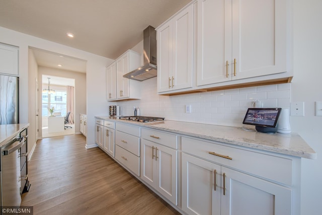 kitchen featuring wall chimney range hood, appliances with stainless steel finishes, white cabinetry, light stone counters, and light wood-type flooring