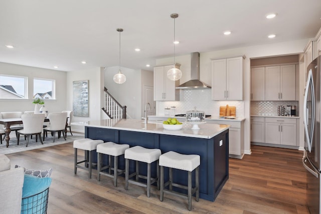 kitchen with decorative light fixtures, stainless steel fridge, dark hardwood / wood-style flooring, a kitchen island with sink, and wall chimney range hood