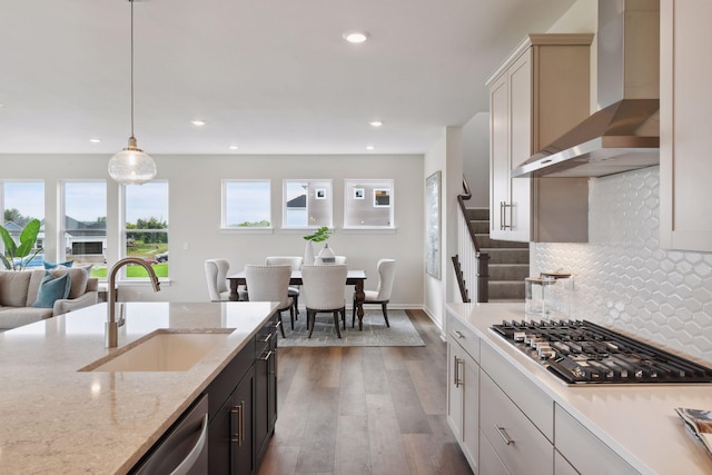 kitchen with sink, appliances with stainless steel finishes, light stone counters, decorative light fixtures, and wall chimney exhaust hood