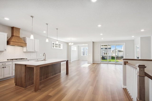 kitchen with premium range hood, an island with sink, sink, white cabinets, and hanging light fixtures