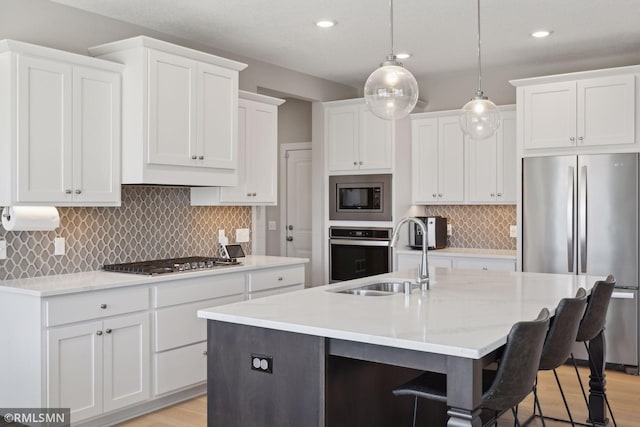 kitchen with a kitchen island with sink, stainless steel appliances, a sink, white cabinets, and pendant lighting