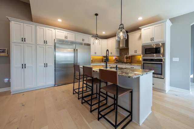 kitchen with stone countertops, decorative light fixtures, white cabinets, built in appliances, and wall chimney exhaust hood