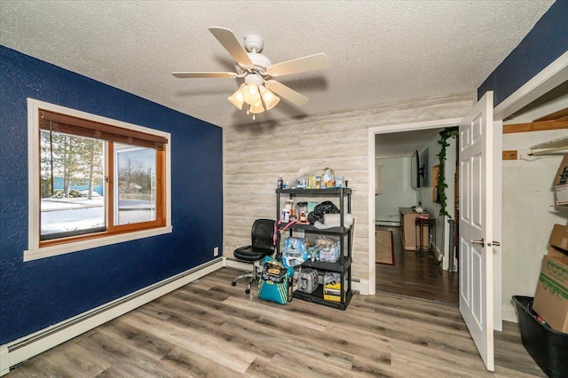 misc room with a baseboard heating unit, a textured ceiling, wooden walls, wood finished floors, and a ceiling fan