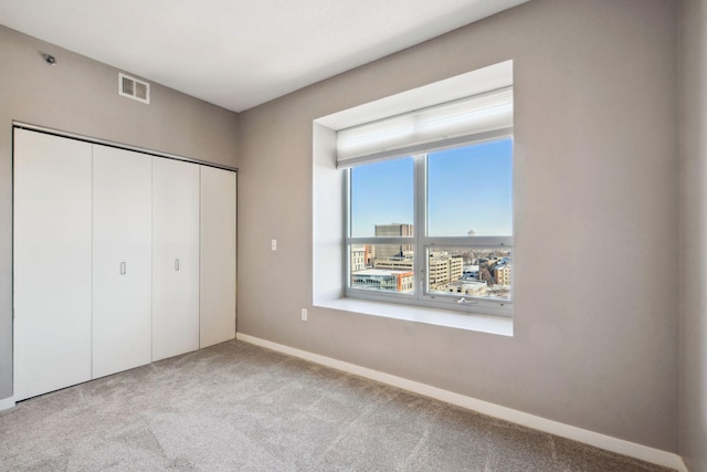 unfurnished bedroom with baseboards, visible vents, light colored carpet, a view of city, and a closet